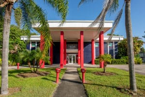 Front view of the Dennis Hernandez Injury Attorneys office in Fort Myers, Florida, with red columns, blue walls, and palm trees lining the entrance.