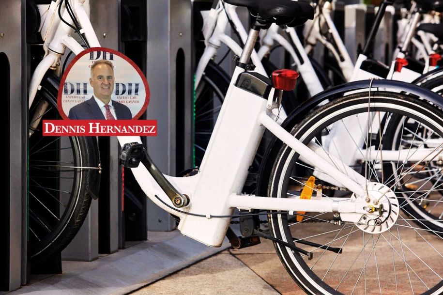 Row of white rental bicycles docked at a bike-share station, with the Dennis Hernandez Injury Attorneys logo overlay