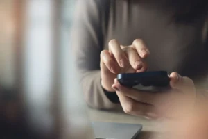Person using a smartphone while seated at a desk, with a laptop nearby and soft lighting in the background.