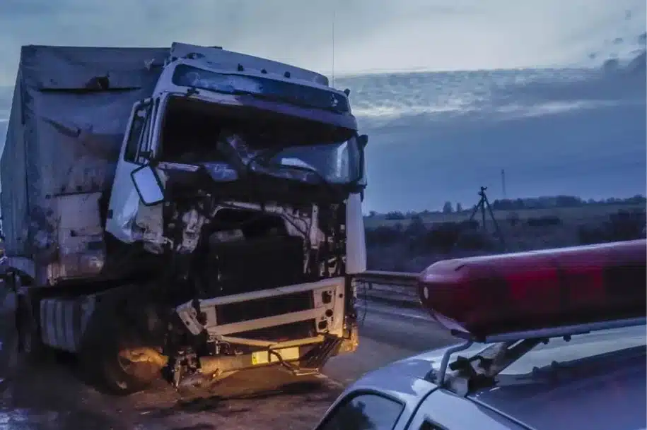 Damaged semi truck at the scene of a highway crash, with a police vehicle and emergency lights nearby.
