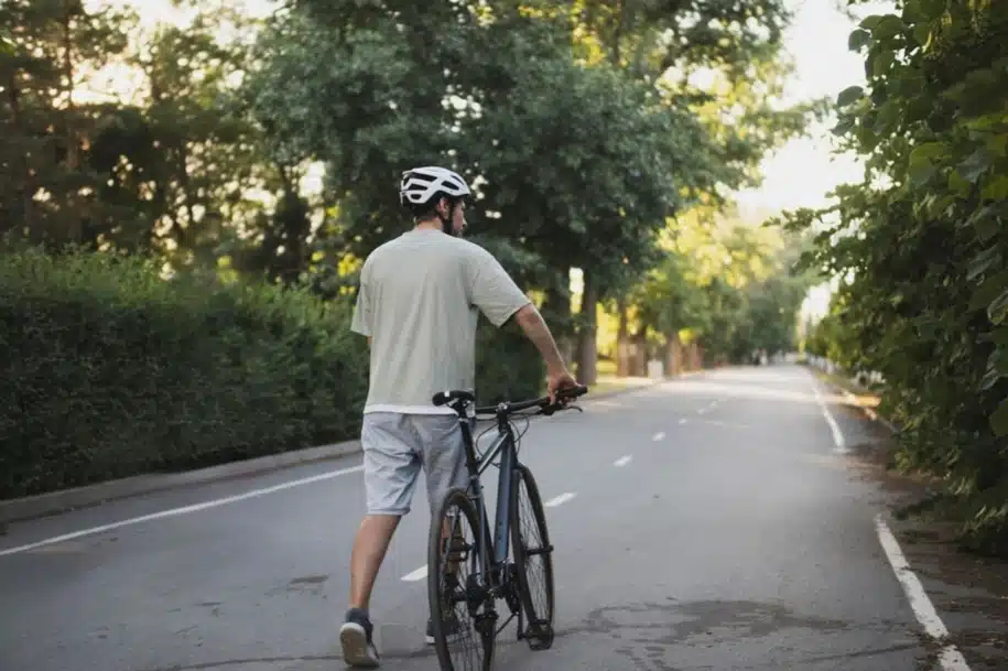 Cyclist wearing a helmet walking a bicycle along a quiet tree-lined road.