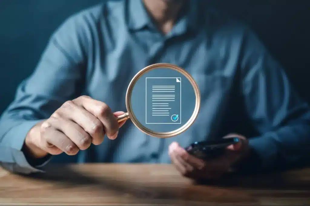 Person holding a magnifying glass over a digital document icon, symbolizing careful review of paperwork.