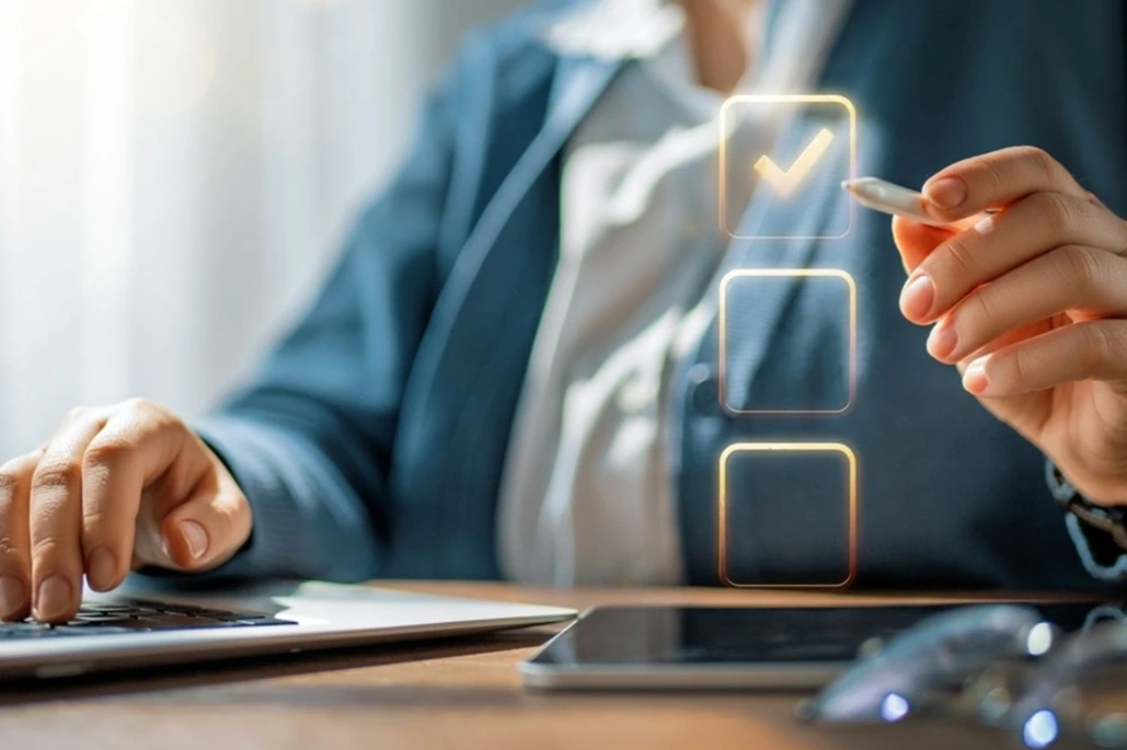 Professional at a desk using a laptop and pen, with a glowing checklist icon showing one box checked.