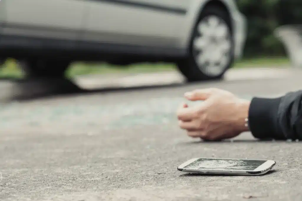 Cracked smartphone on the road near an injured person’s hand, with a car blurred in the background.