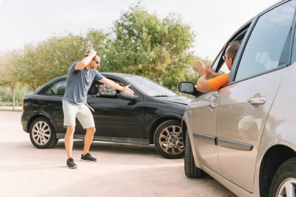 Two drivers arguing after a car accident, one gesturing beside a black car while the other leans out of a silver car window.
