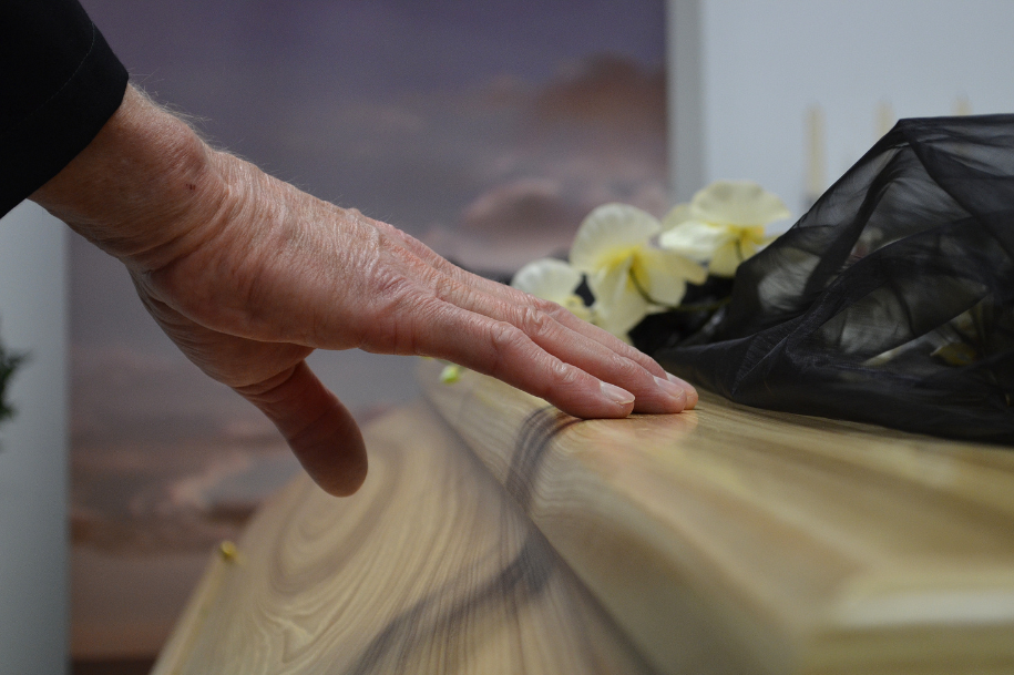 Hand resting on the edge of a wooden casket during a funeral or memorial service.