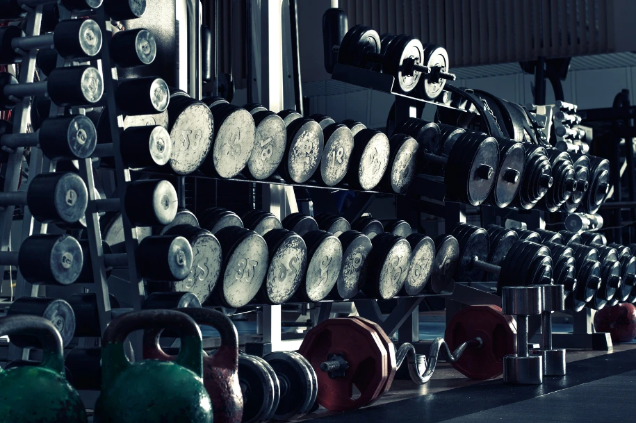 Rows of dumbbells and kettlebells arranged on racks inside a gym weight training area.