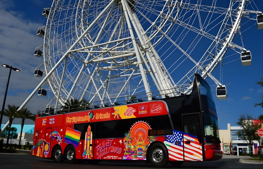 Tour bus parked near a large observation wheel at a busy entertainment district, showing a common tourist transportation area.
