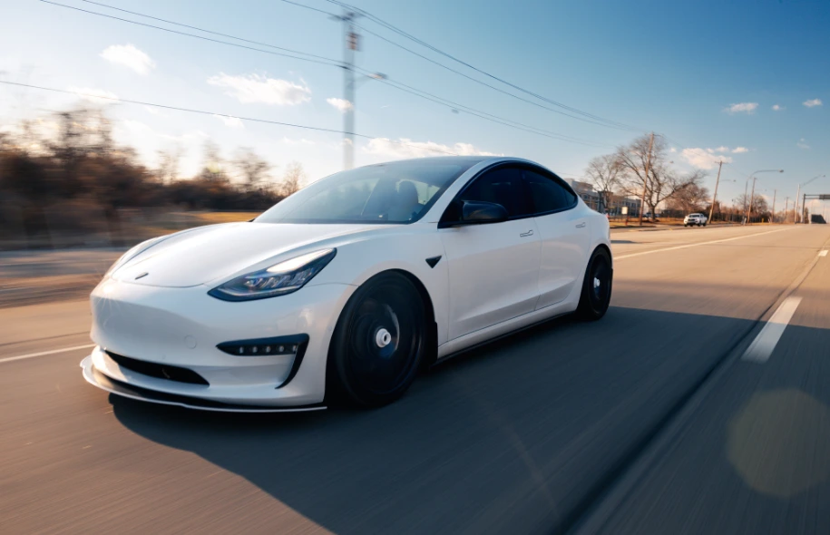 White electric vehicle driving on a roadway during daytime traffic.