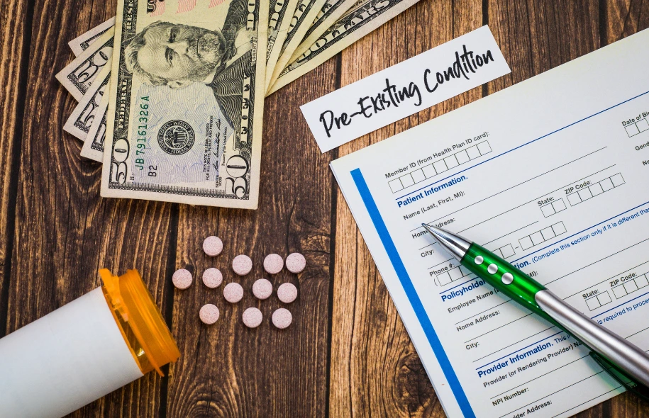 Health insurance form labeled “Pre-Existing Condition” on a wooden table with U.S. dollar bills, prescription pills, a pill bottle, and a pen.