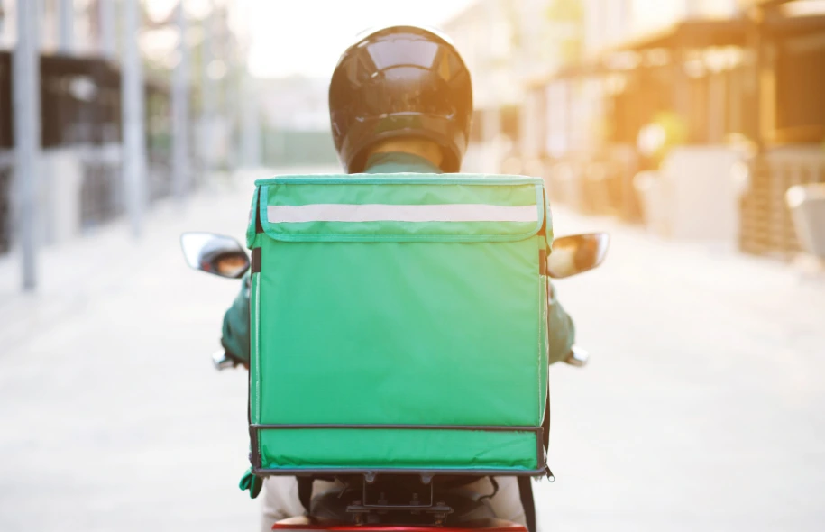Delivery driver wearing a helmet riding a scooter with a green insulated delivery box on the back.