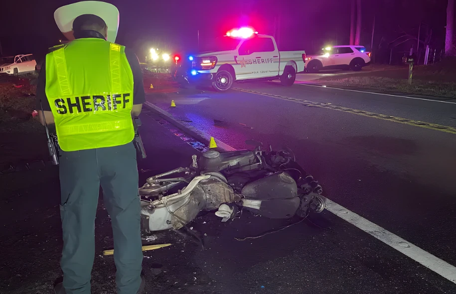 Sheriff’s deputy stands near a damaged motorcycle at a nighttime crash scene with patrol vehicles and emergency lights in the background