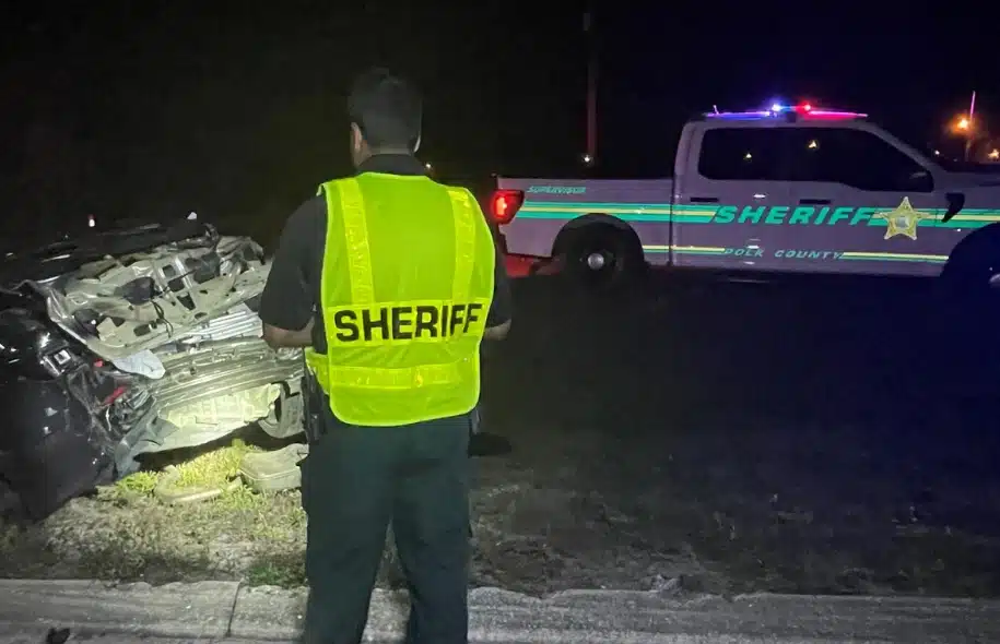Sheriff officer standing near a crashed vehicle with patrol truck lights flashing at night.