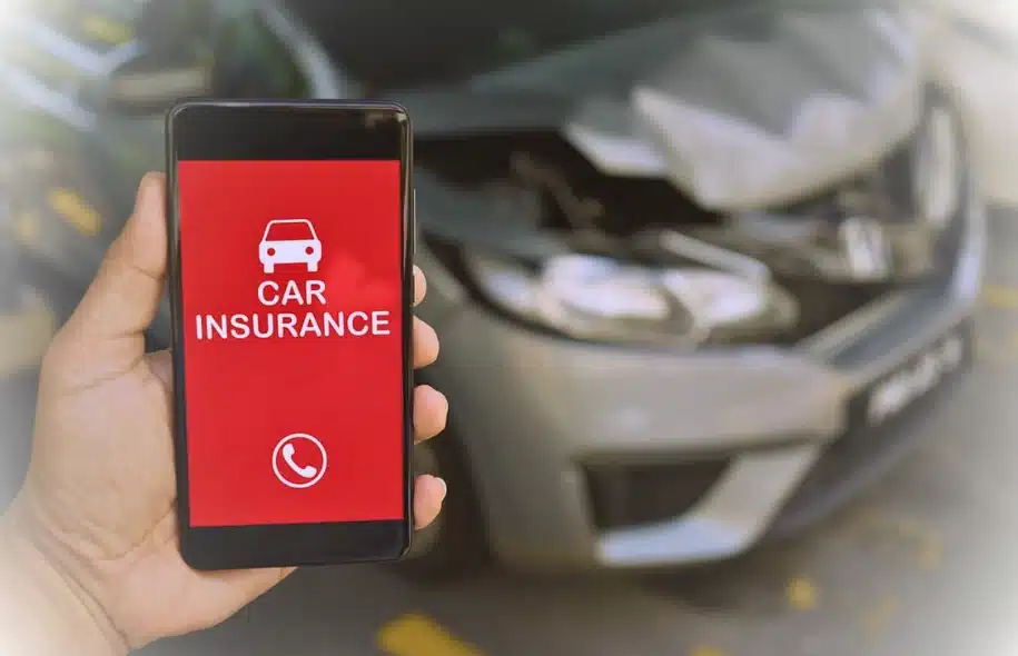 Person holding smartphone displaying car insurance app near damaged vehicle after crash.