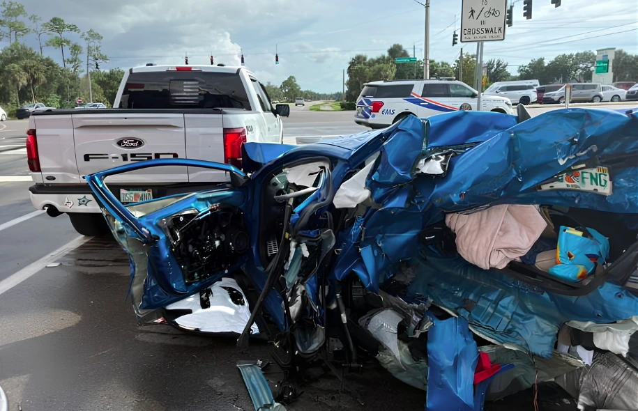 Severely damaged blue car at a Fort Myers crash scene, with a pickup truck and emergency vehicle nearby.